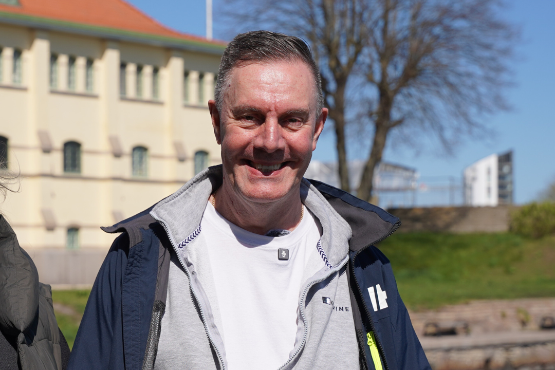 En leende Torgny i ett härligt vårlandskap med byggnad och träd i bakgrunden